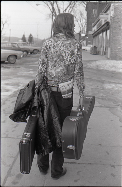 Tom Frank walking away from the camera and             carrying a guitar case, a copy of Free Spirit Press (vol. 1, 4) in his rear pocket, ca. 1973