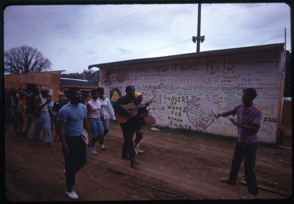 Guitarists leads a line of residents past a graffiti-covered wall in the Resurrection City encampment, ca. June 1968